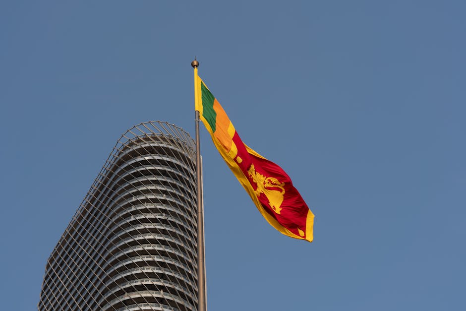 The Sri Lankan flag waving beside a modern Colombo skyscraper against a clear blue sky.